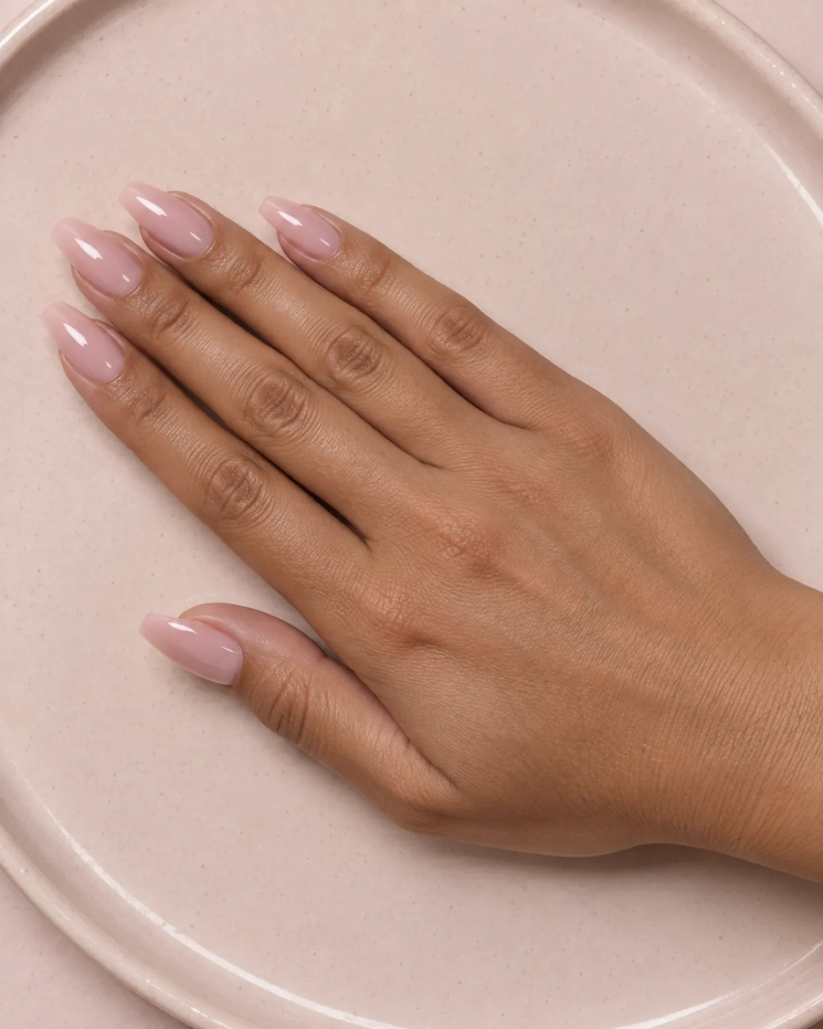 Hand with medium-length coffin nails covered in thick semi-transparent pale pink builder gel with visible apex, resting on a blush ceramic tray