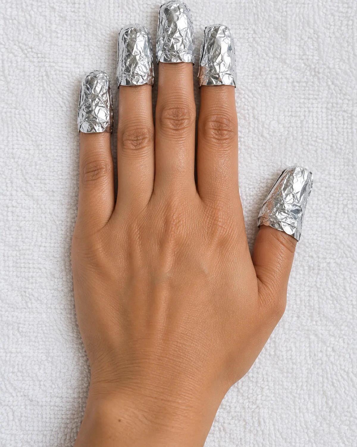 Right hand lying flat on white towel with each fingertip wrapped in a small piece of aluminum foil holding a cotton ball underneath