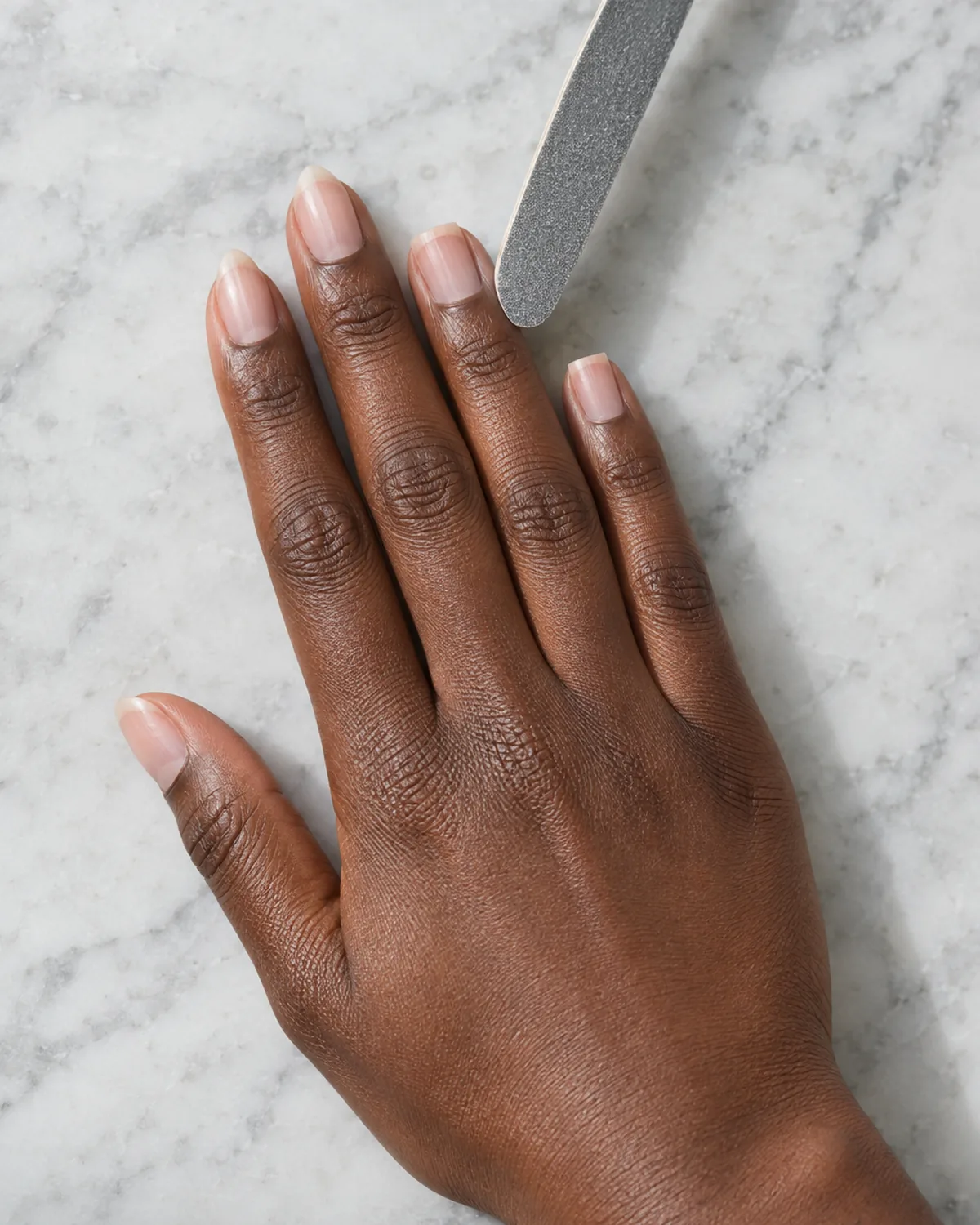 Hand lying flat on grey marble with bare natural nails, a slim grey nail file resting at an angle against the edge of the ring finger nail