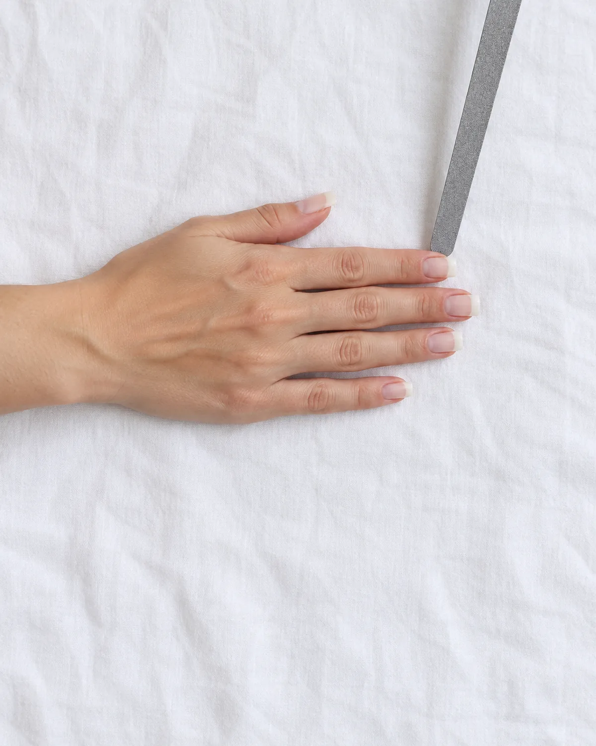 Right hand lying flat on white cotton with bare natural nails filed square with white free edges, a grey nail file resting on the corner of the index nail