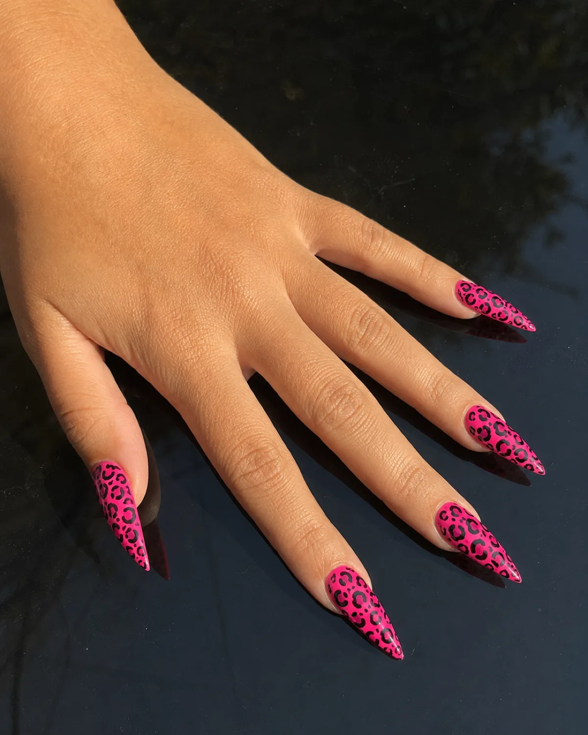 Single left hand with long stiletto nails in neon pink leopard print on glossy black surface