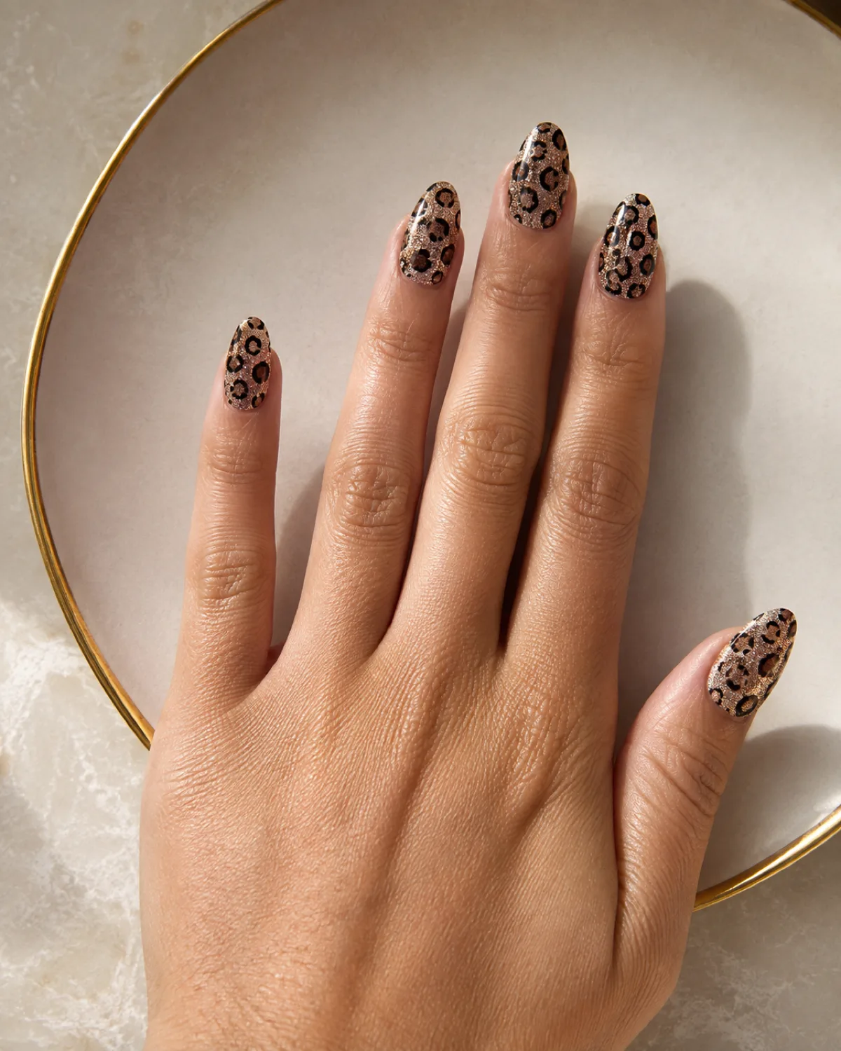Single right hand with medium almond nails in glitter leopard print on gold-rimmed plate