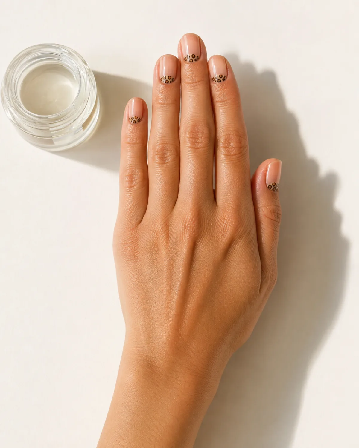Single right hand with short oval nails showing leopard print half-moon design near small glass jar