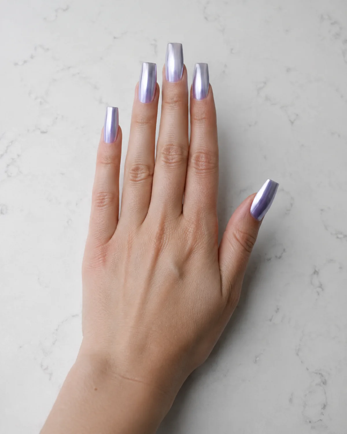Single left hand with long square nails in icy lavender chrome finish resting palm down on white quartz countertop