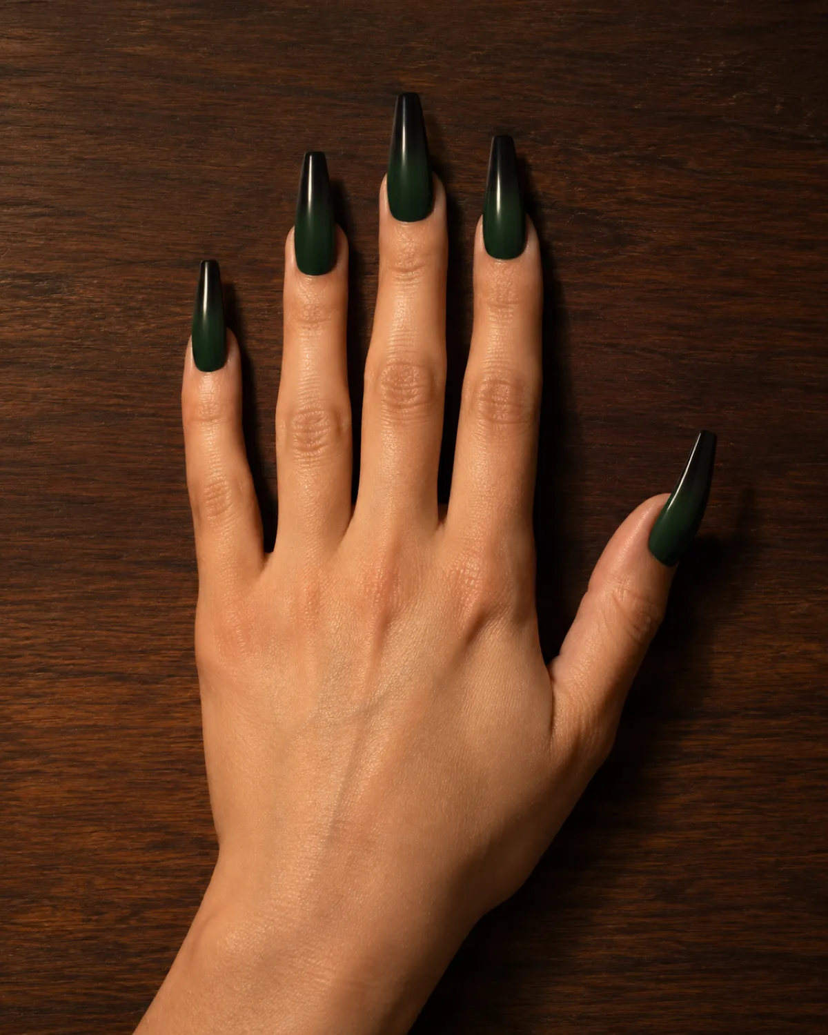 Single left hand with long coffin nails showing emerald-to-black ombré gradient resting palm down on dark walnut table