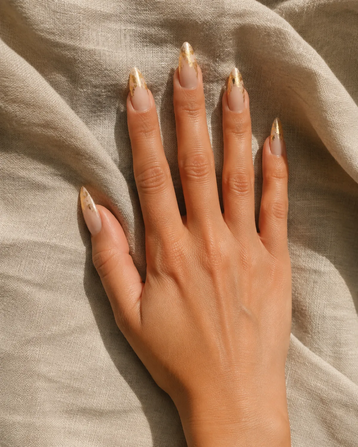 Single right hand with long almond nails showing warm nude base with gold-leaf foil accents resting palm down on raw linen