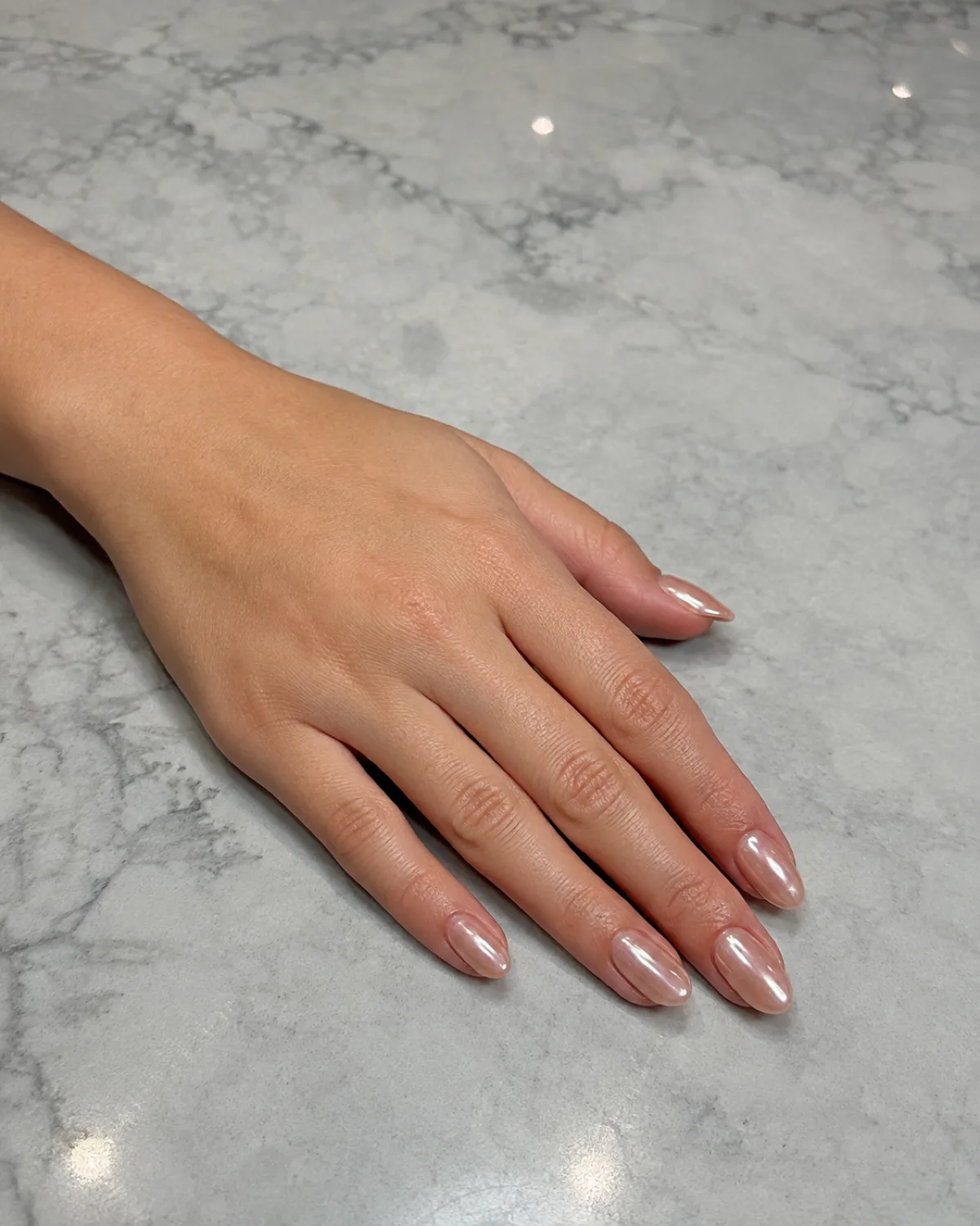 Single left hand with medium oval nails in warm pearl chrome glazed-donut finish resting palm down on grey marble counter