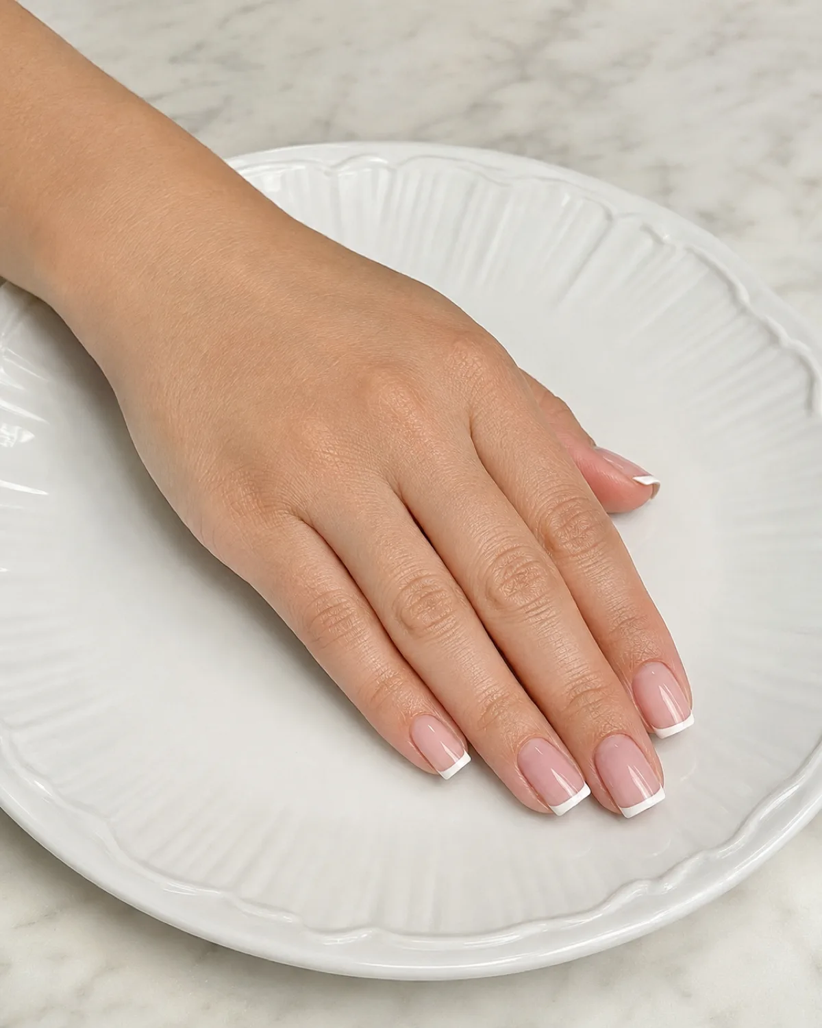 Single left hand with medium square nails showing sheer pink base and crisp white micro-French tips resting on white ceramic plate