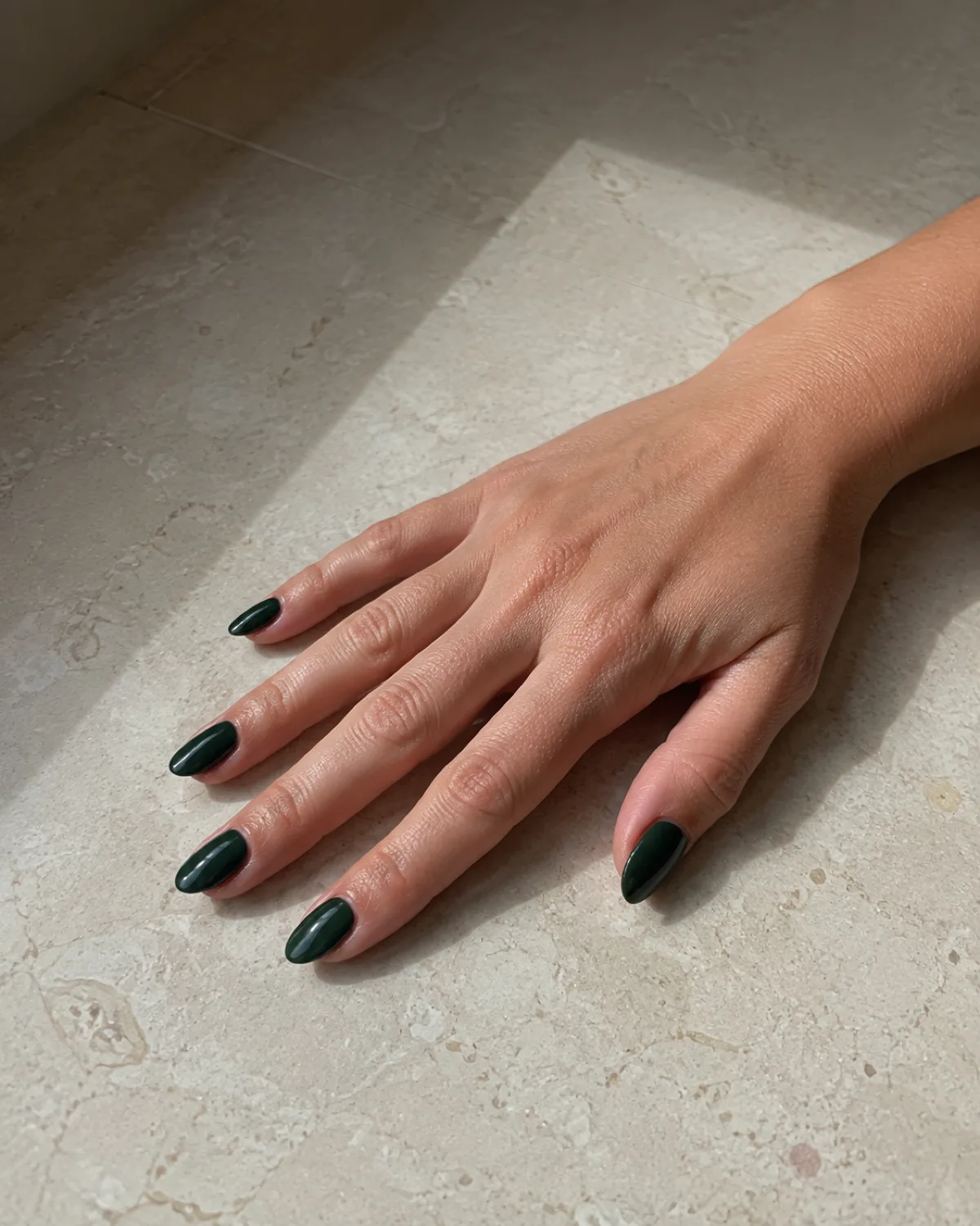 Single right hand with medium almond nails in deep forest green crème gloss resting flat on cream stone tile with fingers fanned