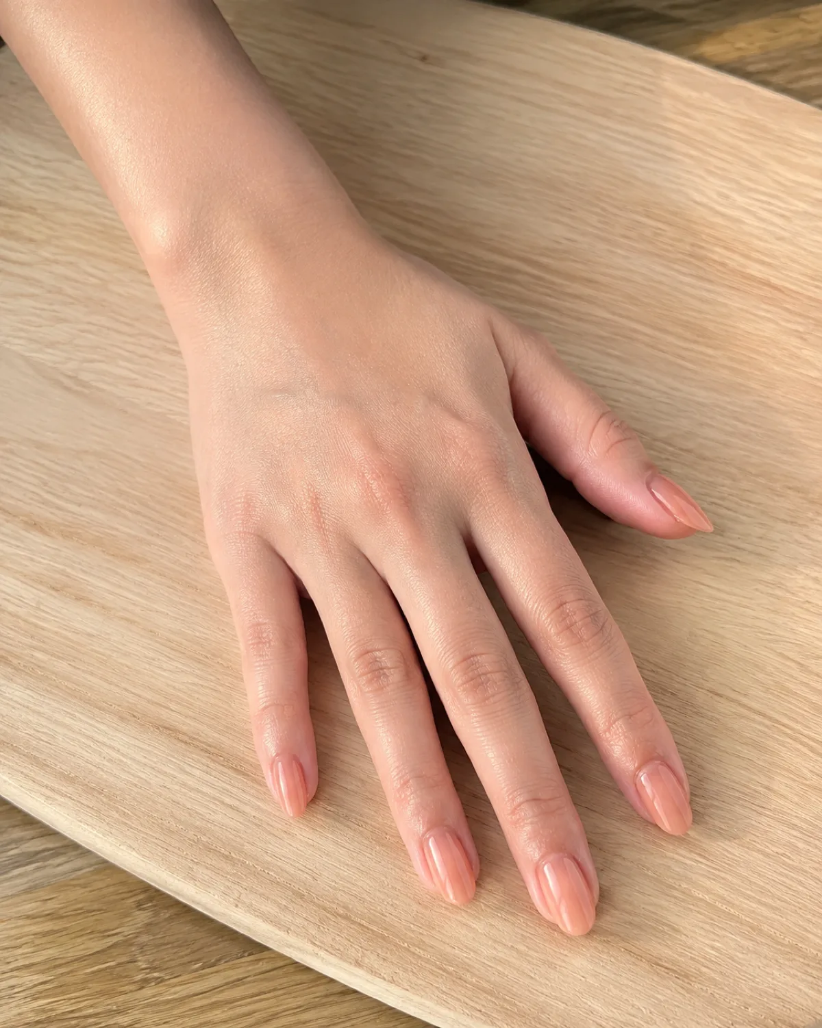 Single left hand with medium oval nails in sheer translucent warm peach jelly finish resting palm down on light wood cutting board