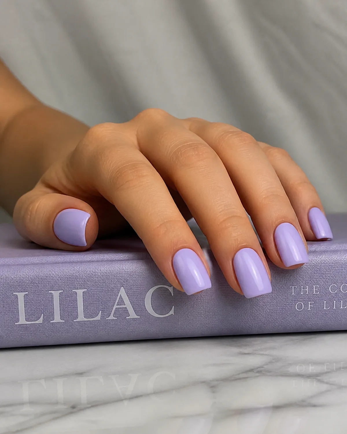 Single left hand with medium square nails in pale lavender crème gloss resting on a lilac book spine with fingers slightly curled