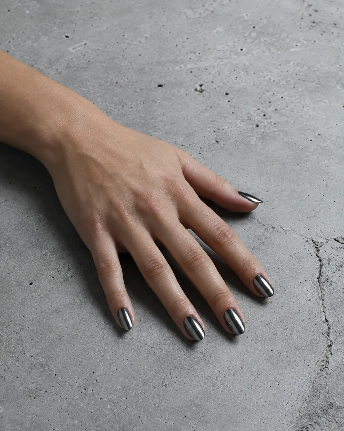 Single left hand with medium oval nails in gunmetal pewter metallic finish resting on raw concrete surface with fingers naturally spread