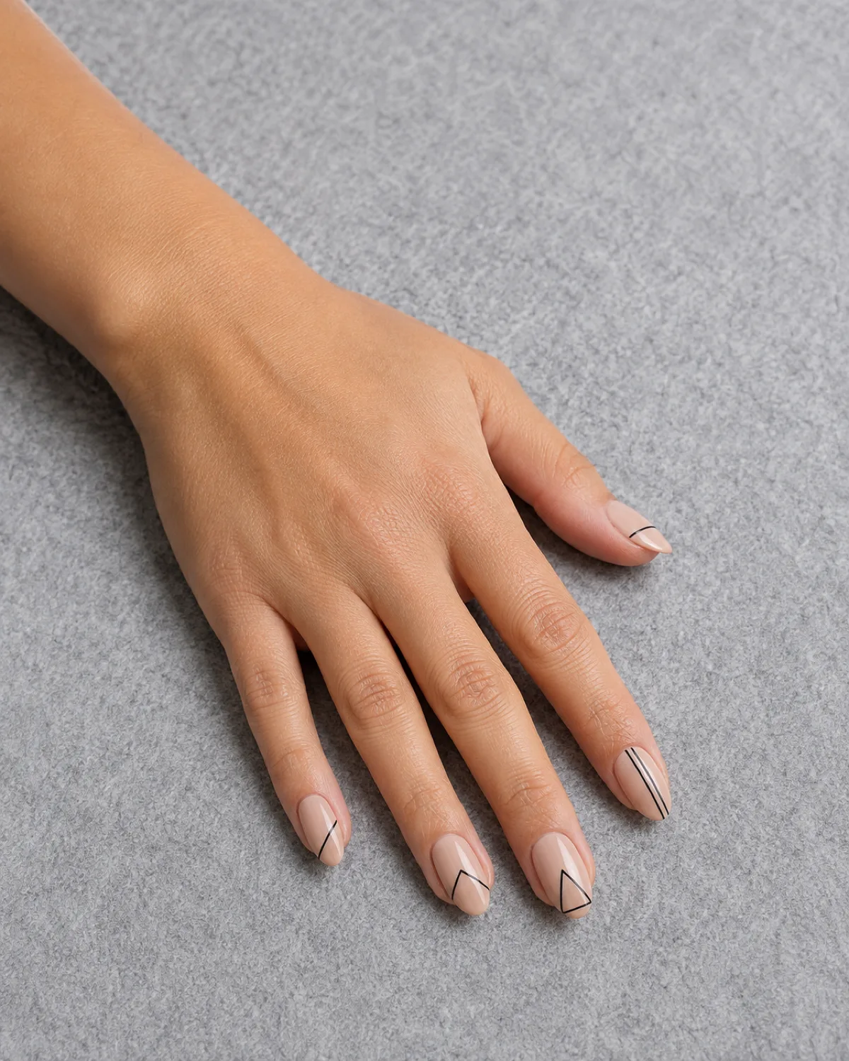 Single left hand with medium almond nails showing warm nude base with single thin black geometric lines resting palm down on light grey felt