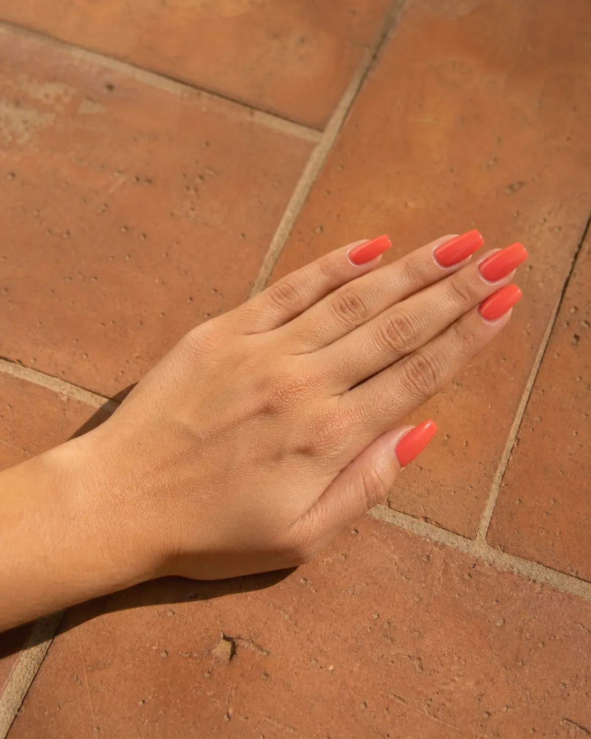 Single left hand with medium coffin nails in bright warm coral high gloss crème resting flat on terracotta tile with fingers together