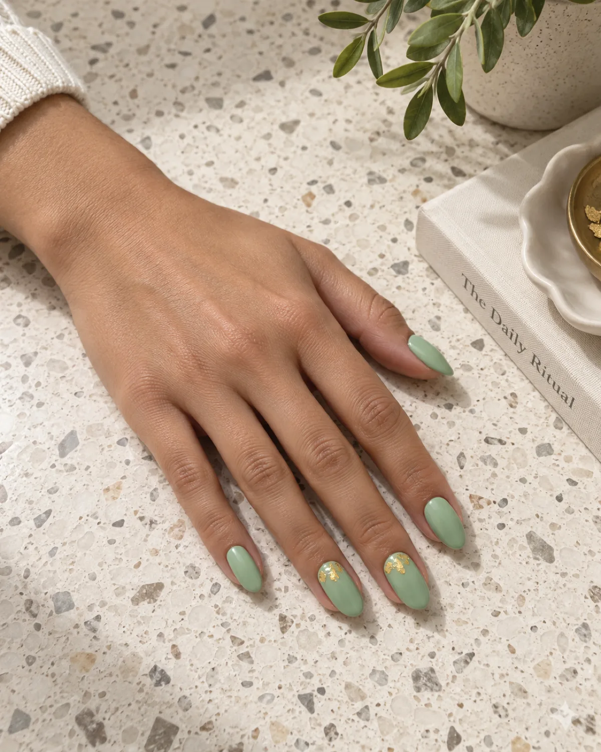 Mint green nails with gold leaf flakes near cuticle on almond shape on cream terrazzo surface