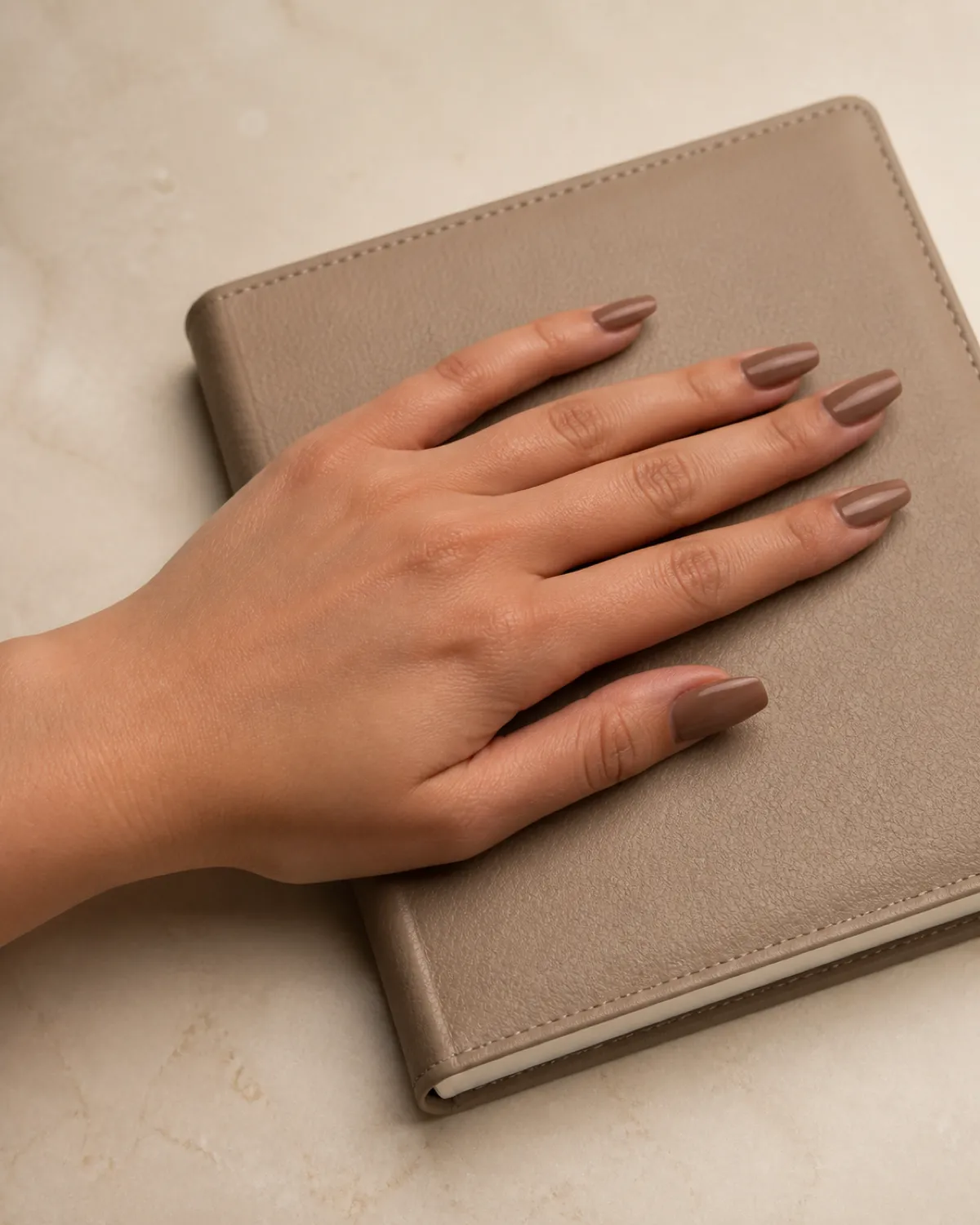 Single left hand with medium coffin nails in café au lait gloss resting palm down on beige leather journal