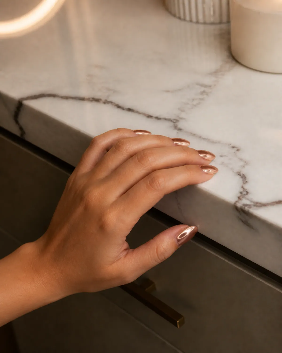 Single left hand with medium almond nails in rose gold chrome on nude base angled from bottom-left corner with fingers draped over marble vanity edge