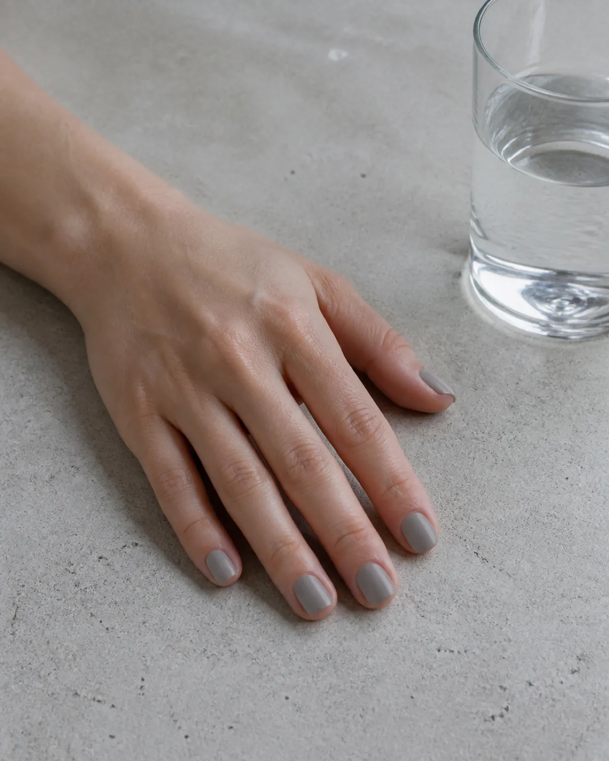Single left hand with short square nails in dove grey matte resting palm down on grey concrete beside glass of water
