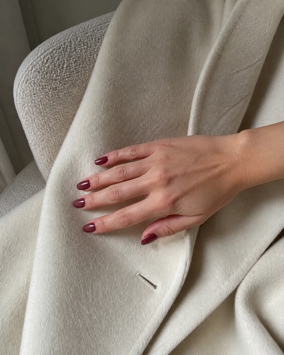 Single right hand with medium oval nails in sheer translucent bordeaux resting palm down on cream wool coat