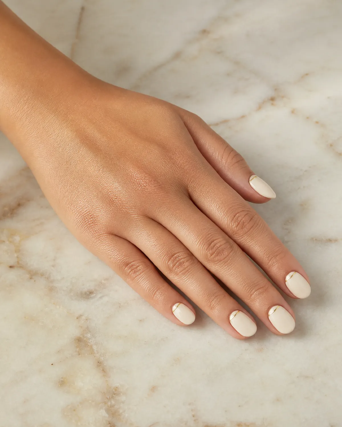 Single left hand with short almond nails in warm ivory and micro gold line at cuticle resting palm down on cream marble