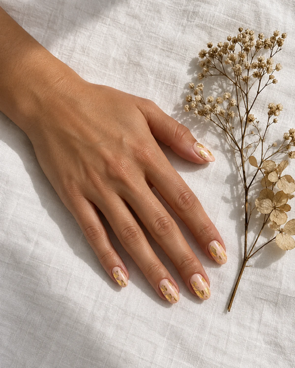 Single left hand with creamy peach oval nails with gold leaf flecks, alongside a dried botanical on white linen