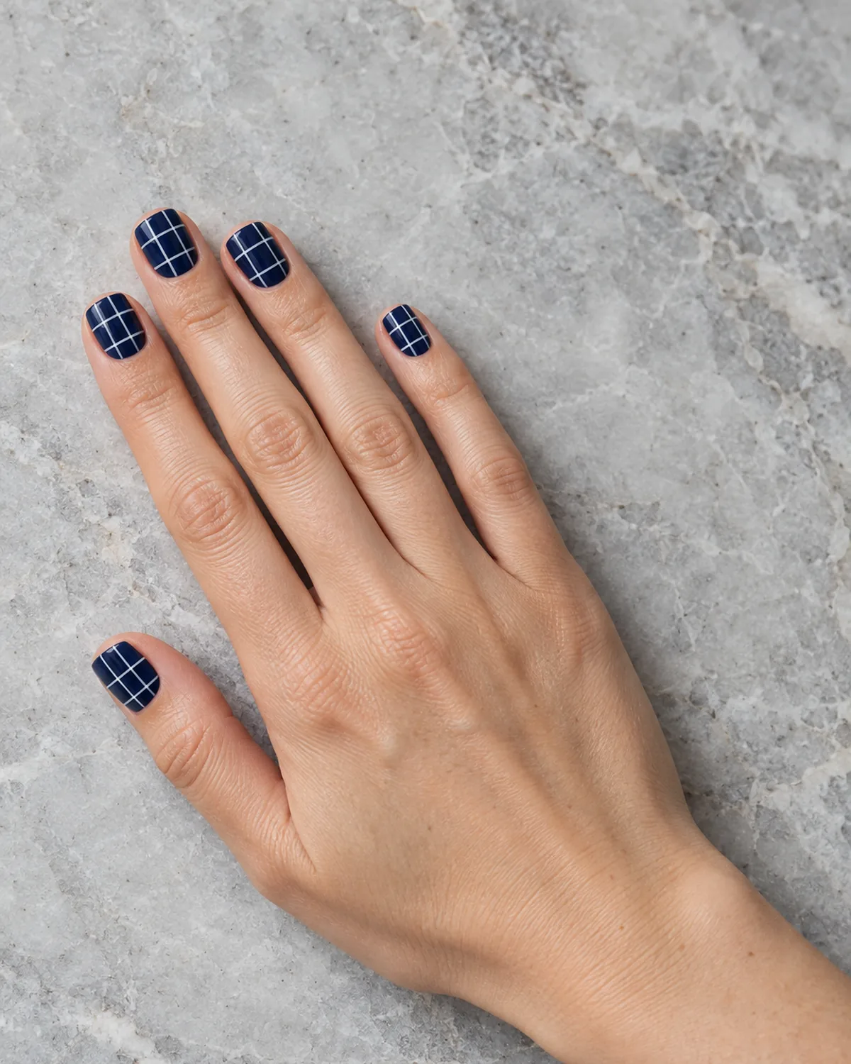 Hand lying on grey stone with navy windowpane check nails with thin white grid lines on short square shape