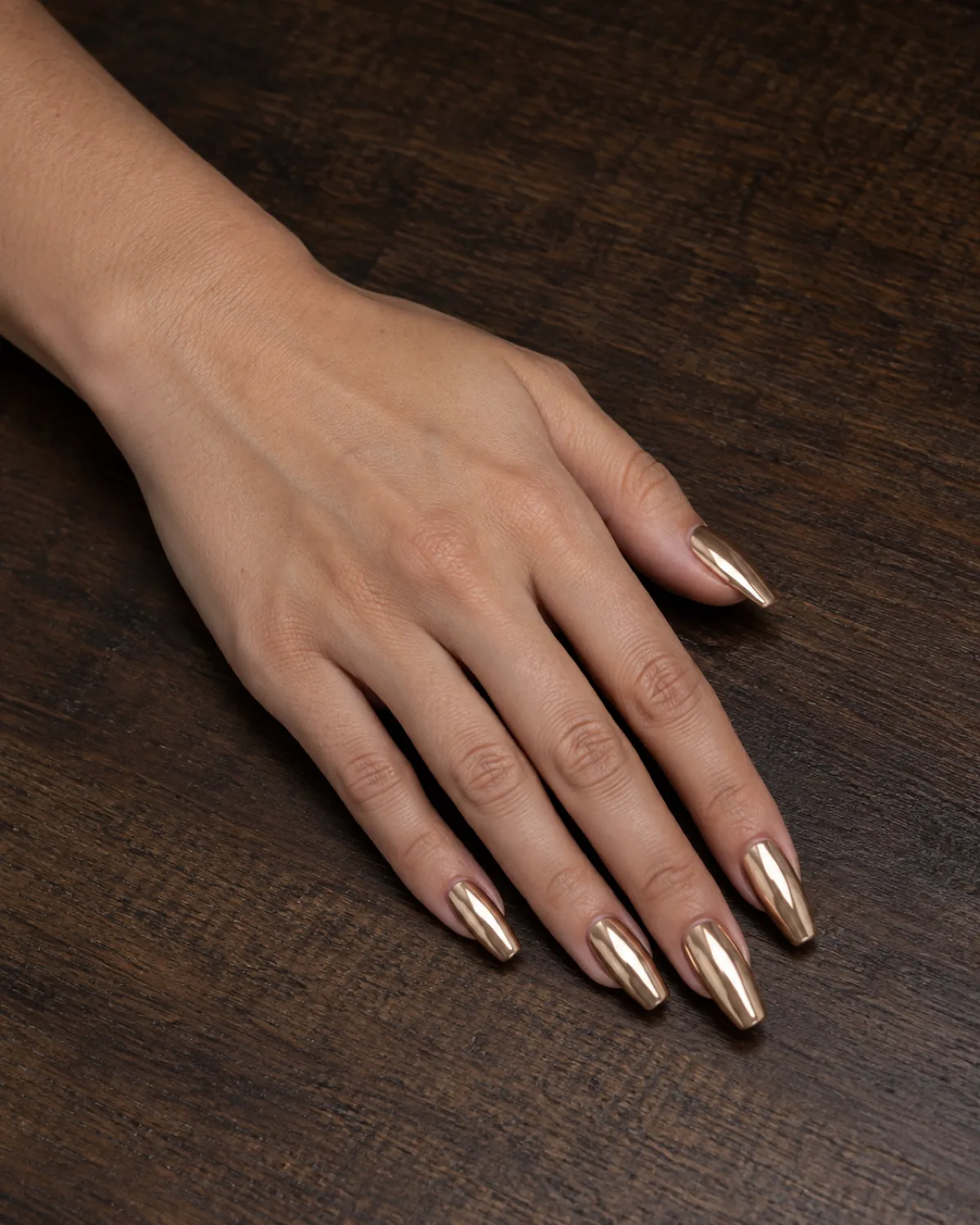 Single left hand with coffin nails in champagne chrome mirror finish resting palm down on dark wood surface