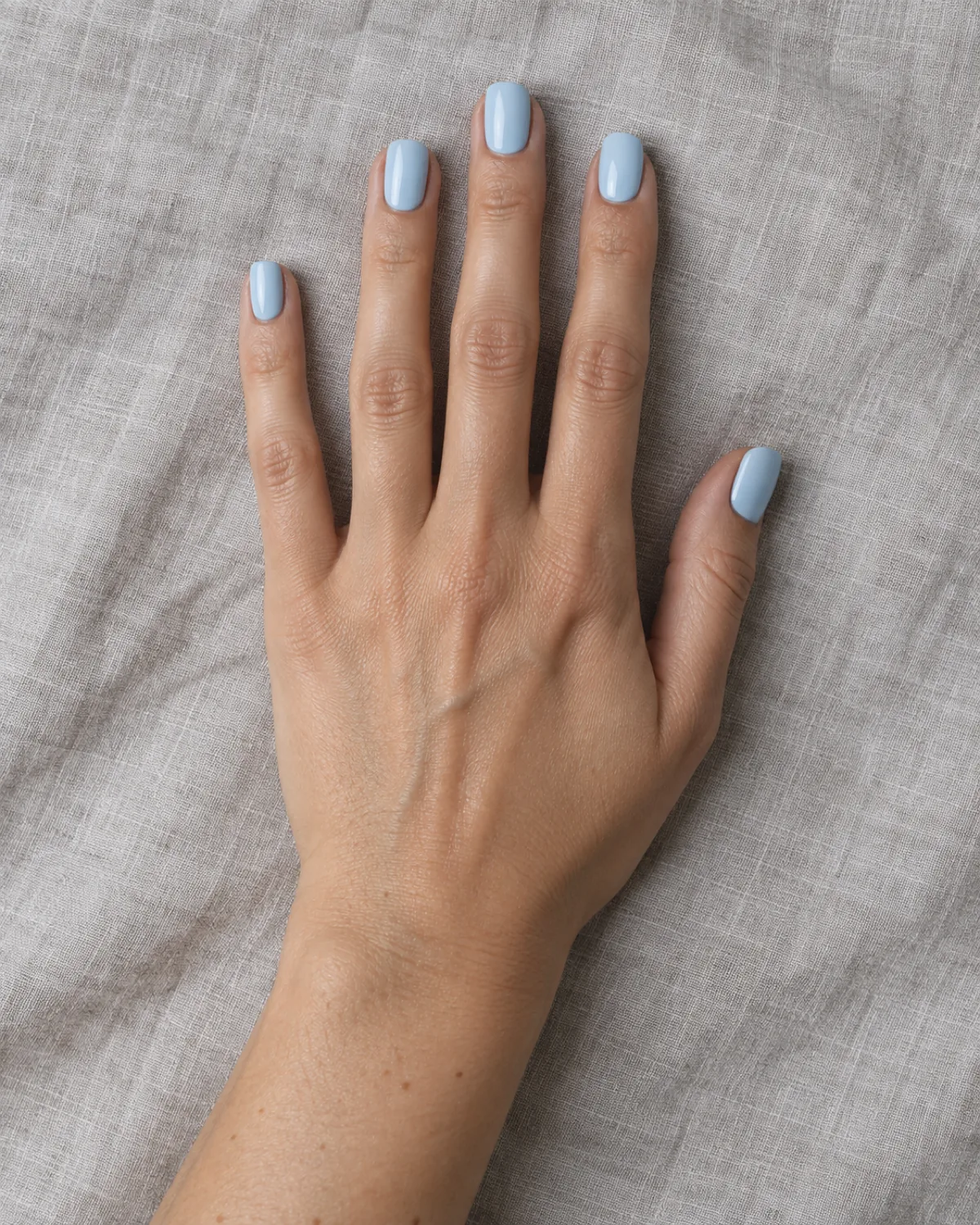 Single right hand with short square nails in pastel baby blue crème polish resting palm down on light grey linen