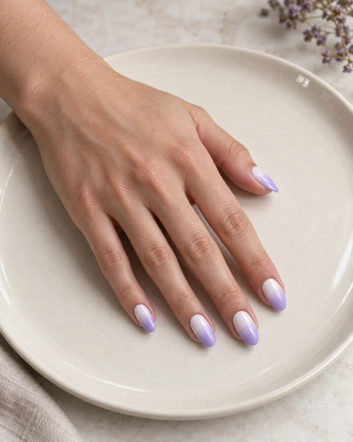 Single left hand with almond nails in white-to-lavender ombré gradient resting palm down on cream ceramic plate