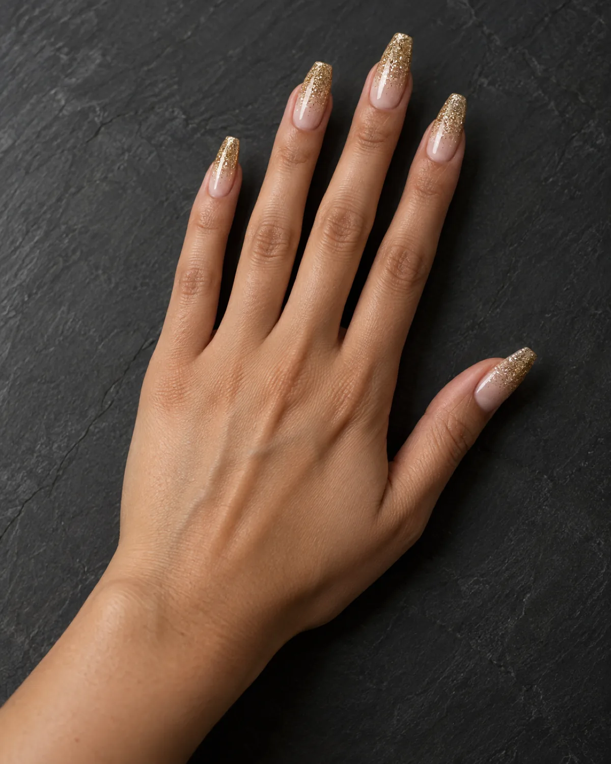 Single right hand with coffin nails in nude base and gold glitter gradient at tips resting palm down on dark slate