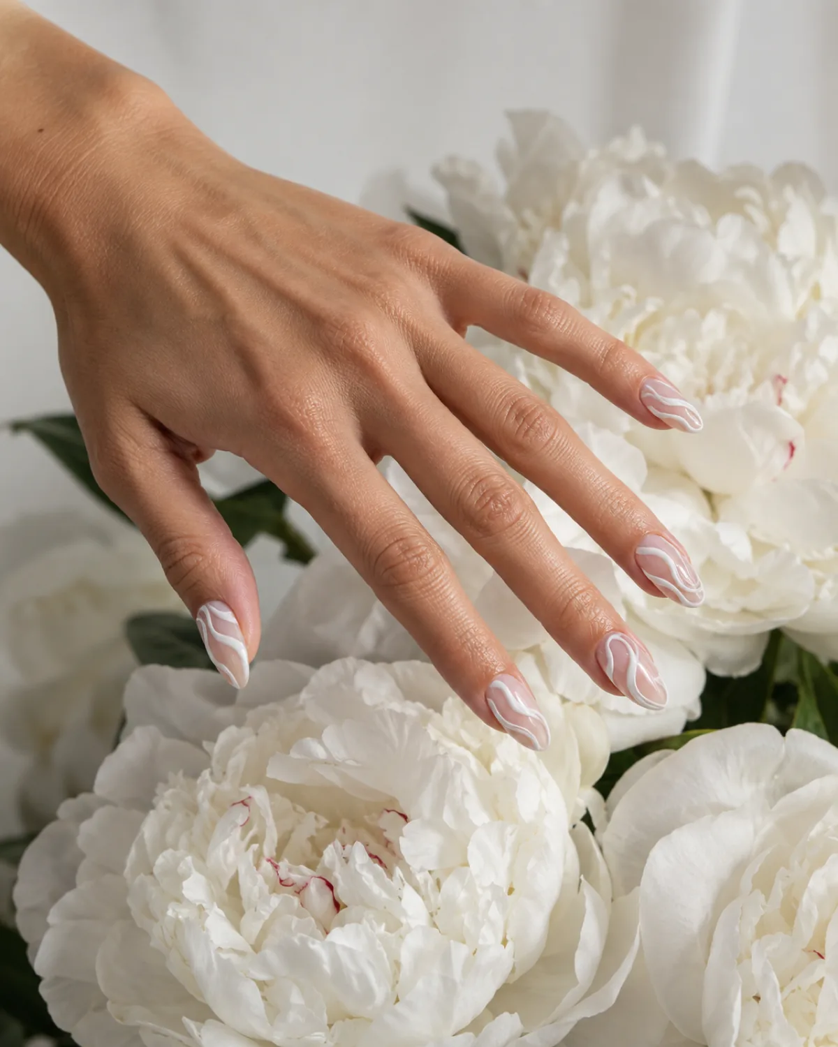 Single left hand with almond nails in sheer pink base and raised white swirl line art hovering above white peony flowers