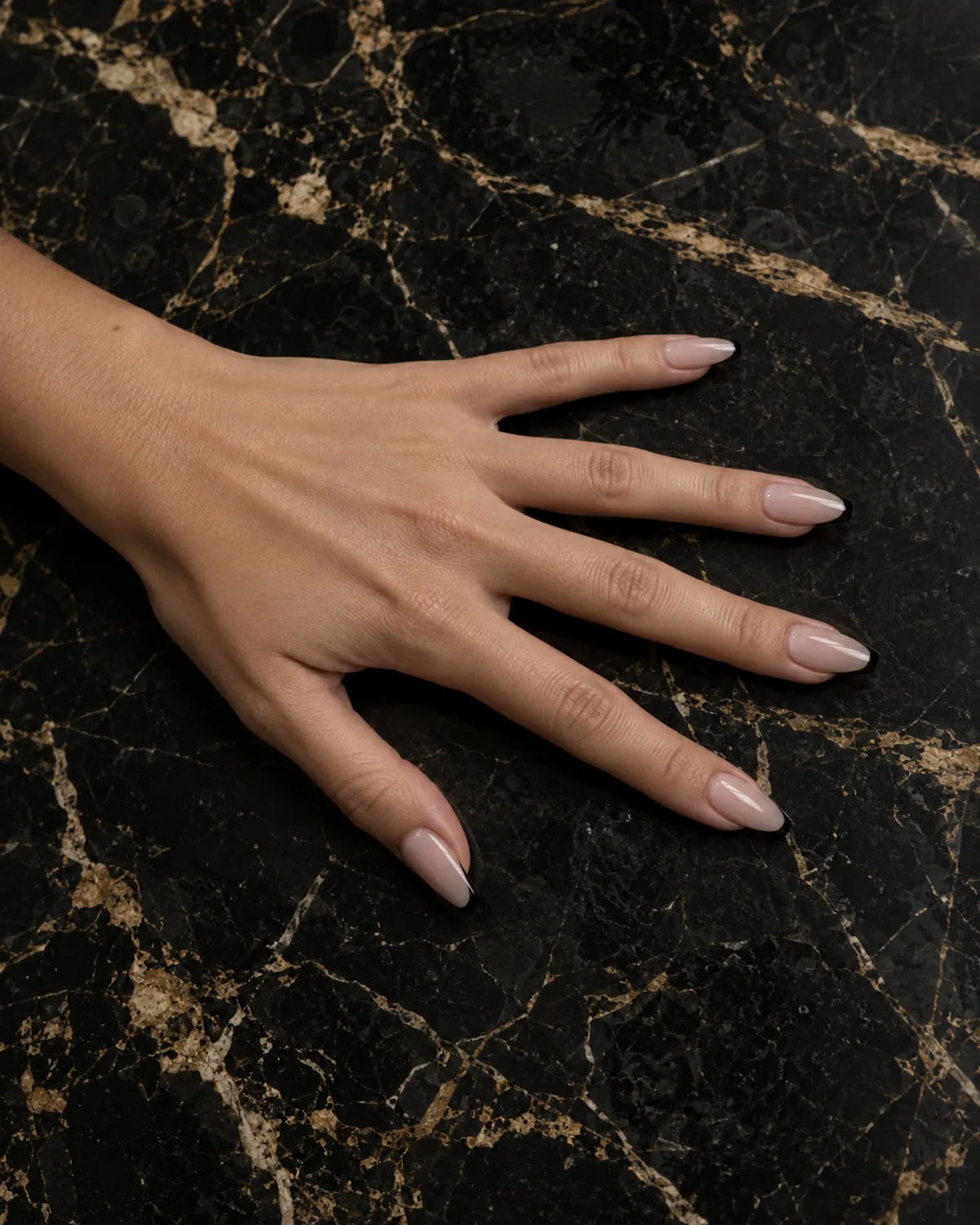 Single left hand with coffin nails in nude sheer base and thin black micro-French tips resting palm down on black marble with gold veining