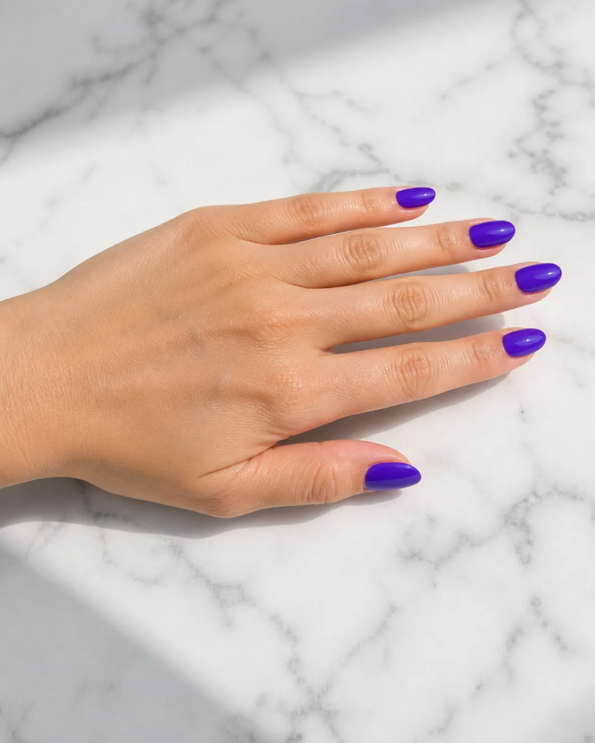 Single left hand with medium almond nails in royal purple crème gloss resting palm down on white marble