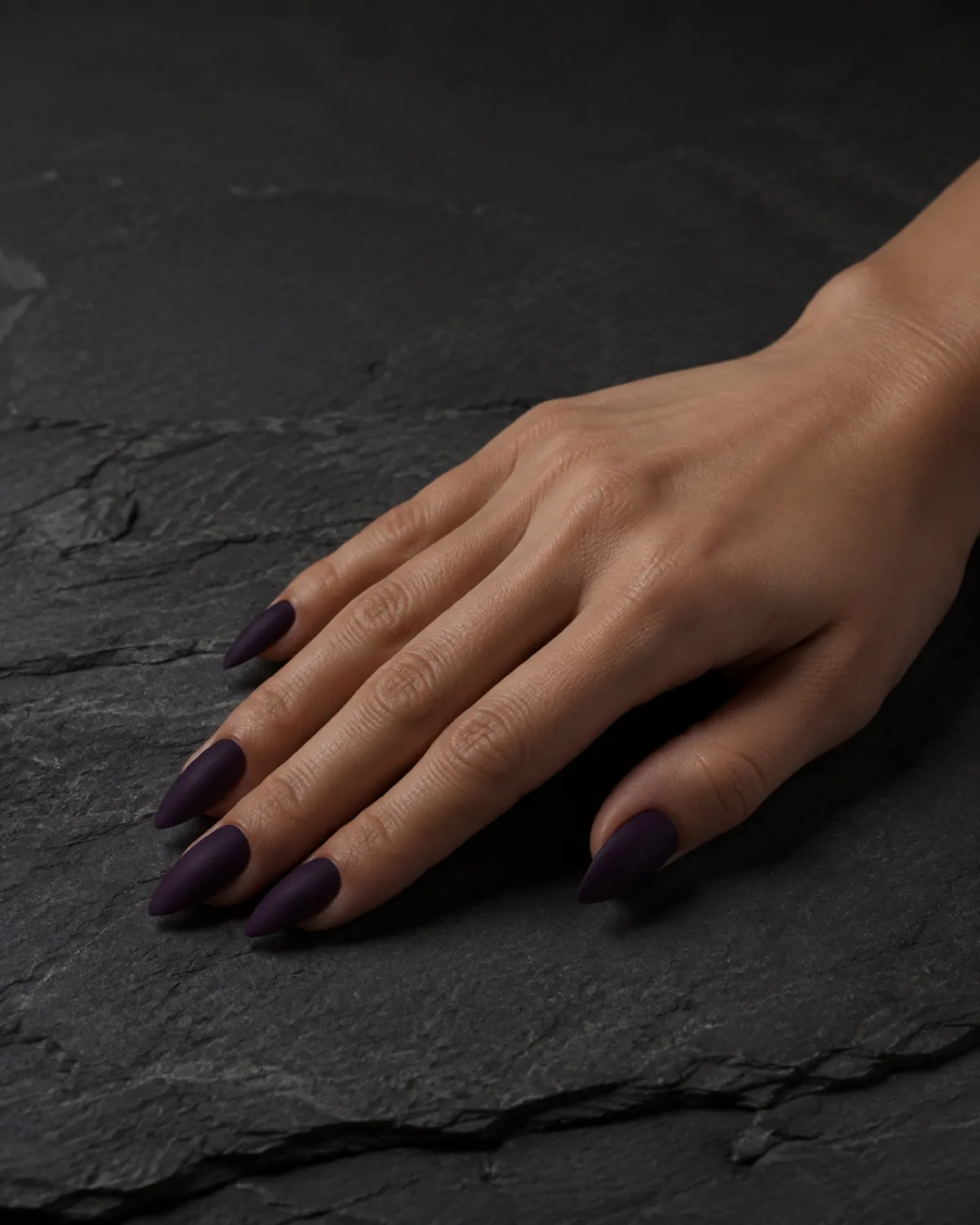 Single right hand with medium stiletto nails in eggplant matte resting palm down on dark slate
