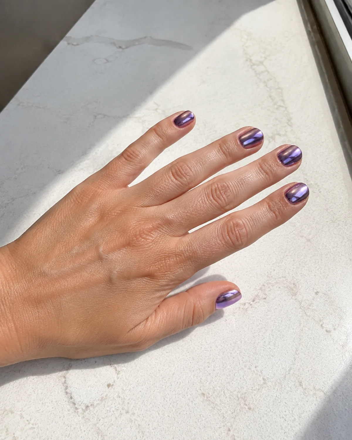 Single left hand with short squoval nails in amethyst chrome mirror finish catching light on white quartz counter