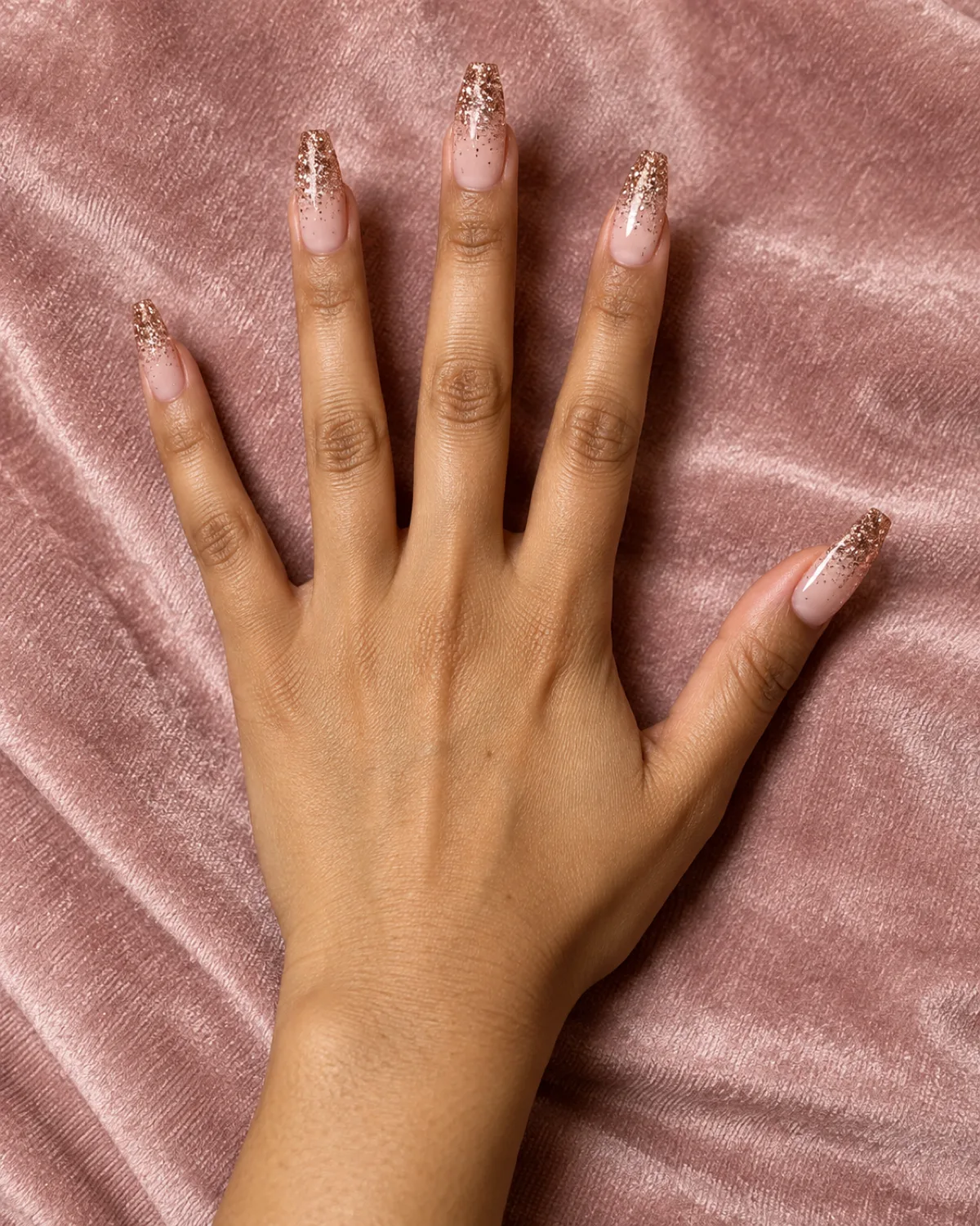 Single right hand with coffin nails showing rose gold glitter gradient on blush pink velvet fabric