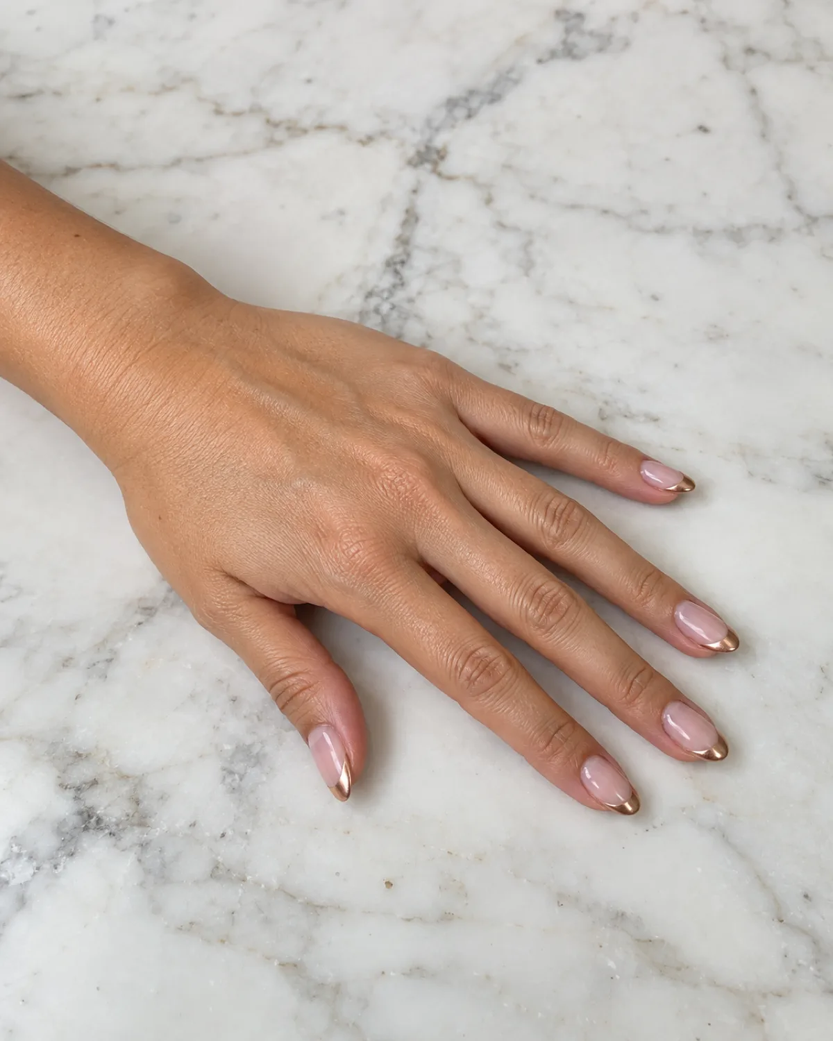 Single left hand with oval nails showing rose gold chrome French tips on white marble countertop