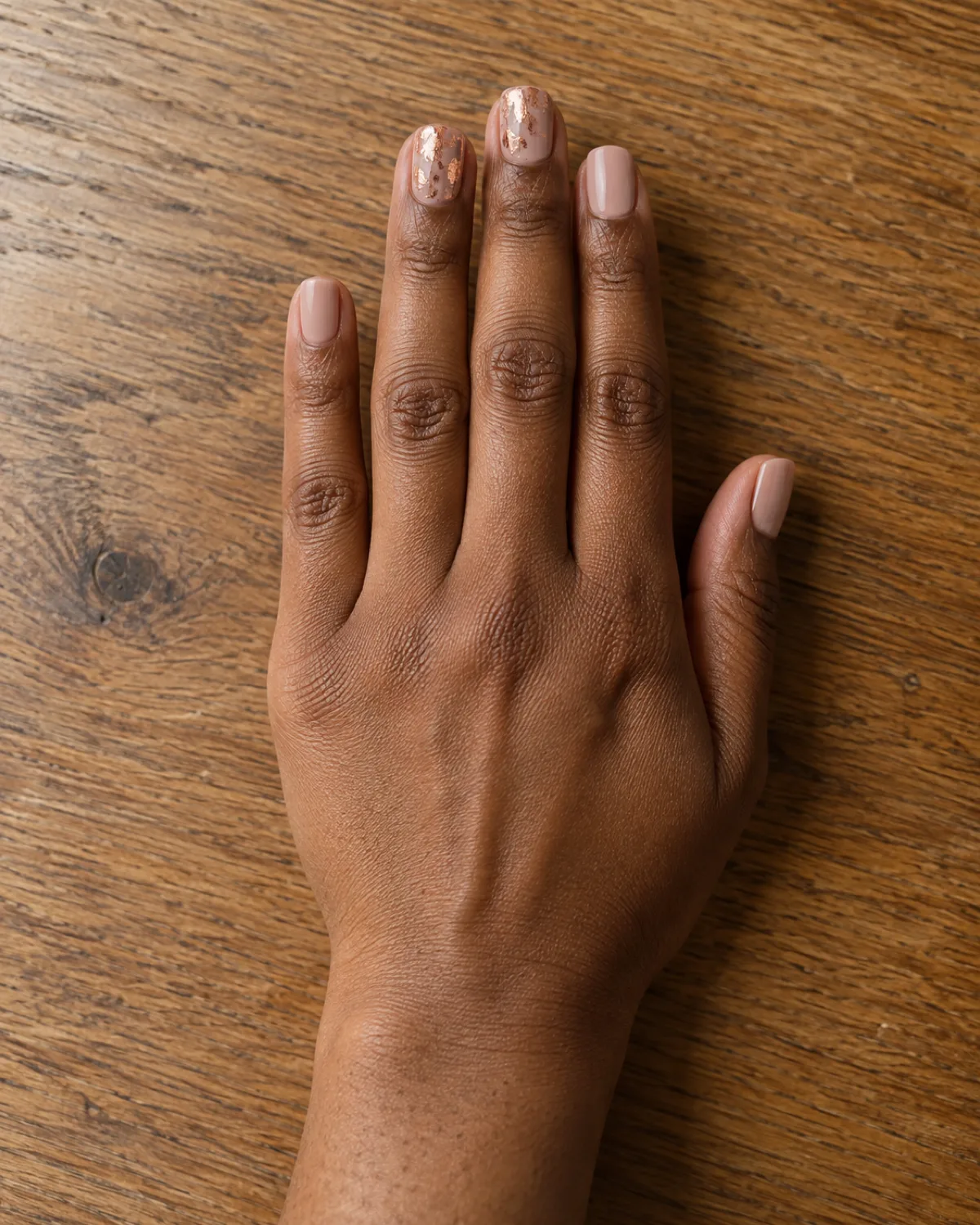 Single right hand with short square nails showing rose gold foil leaf accent on warm oak wood tabletop