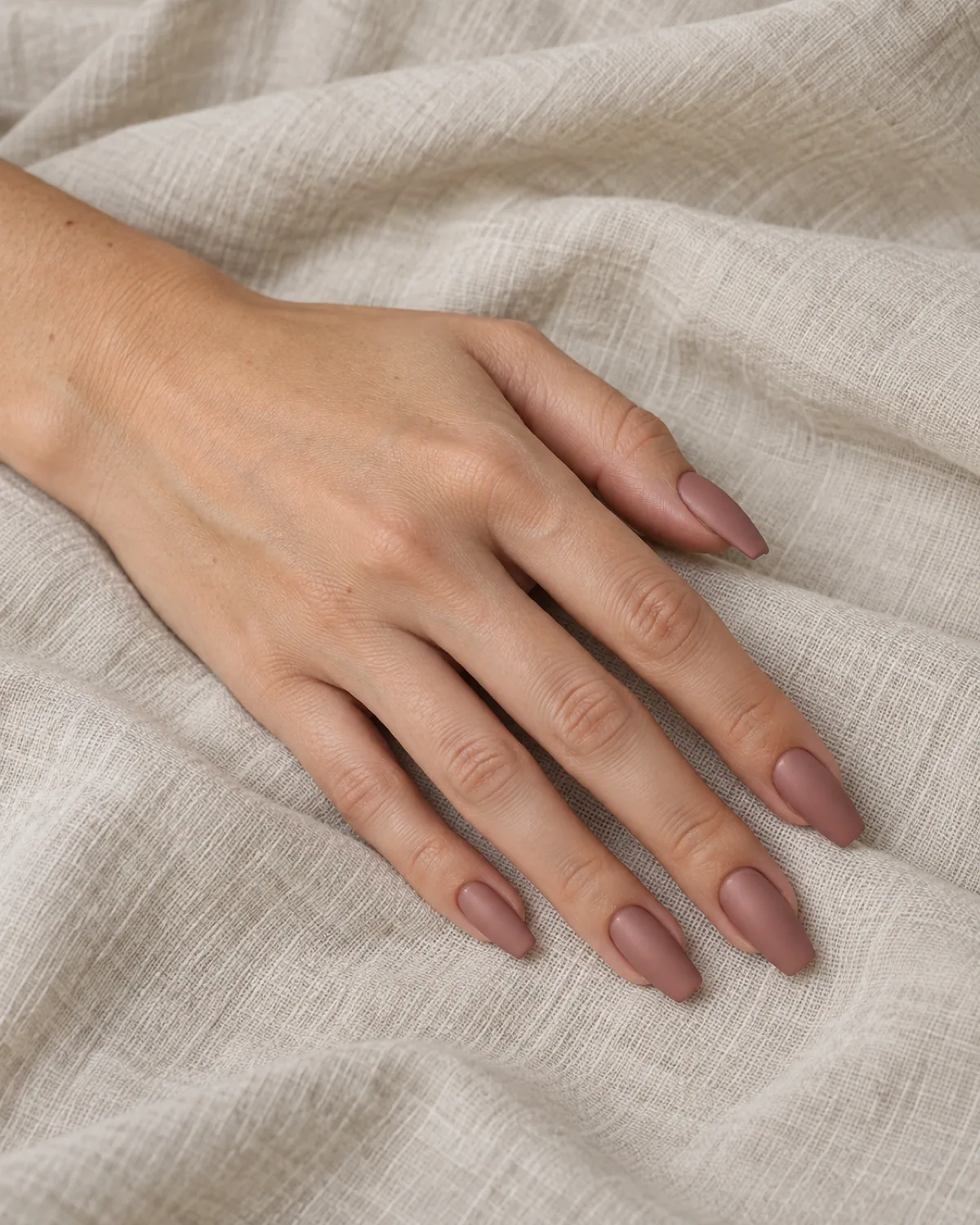 Single left hand with coffin nails in opaque matte dusty rose on cream linen cloth