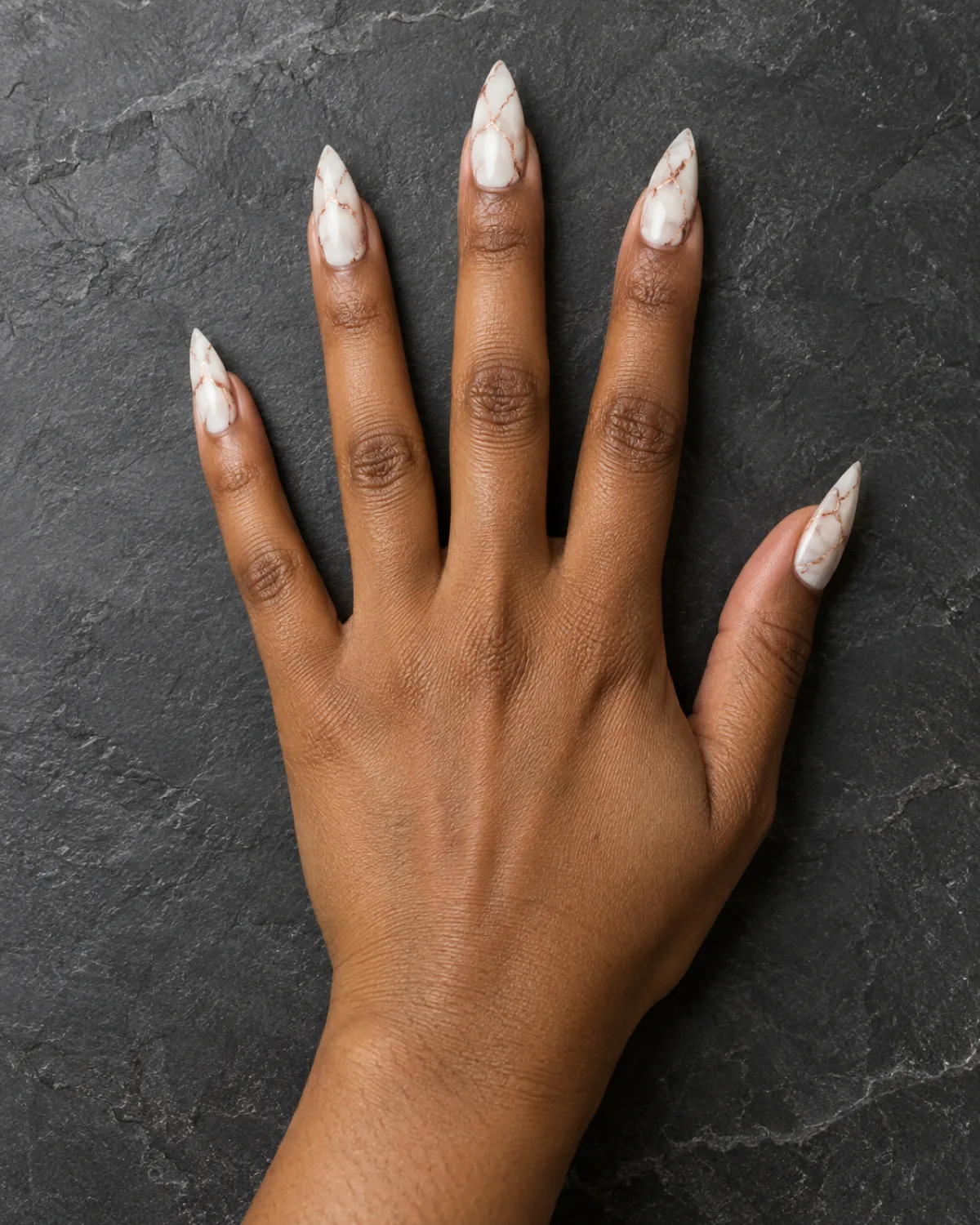 Single right hand with stiletto nails showing white marble effect with rose gold veining on dark grey slate surface
