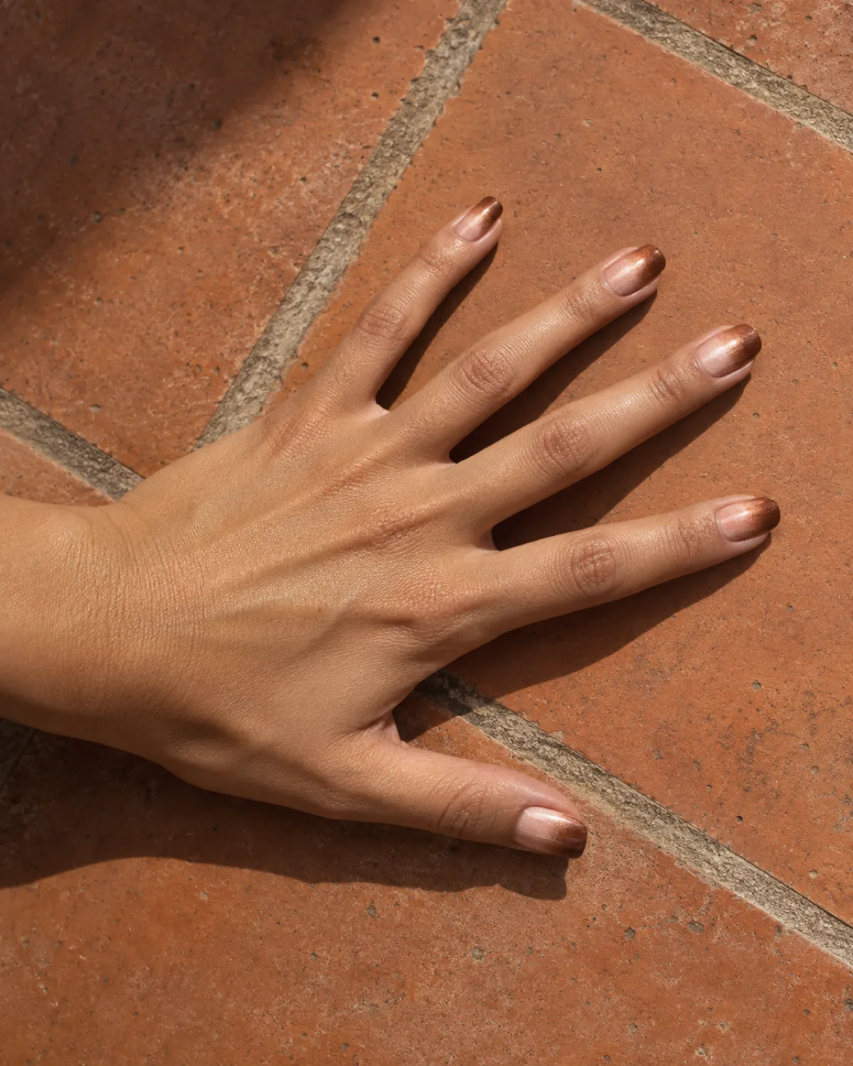 Single left hand with squoval nails showing ombré gradient from rose gold to copper on terracotta tile surface