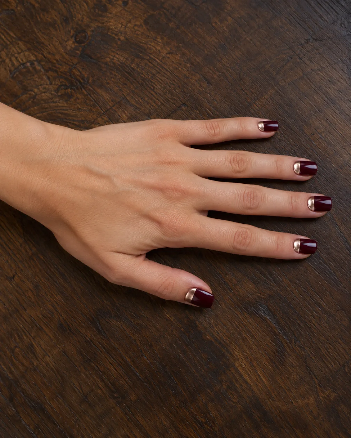 Single left hand with square nails showing burgundy crème with rose gold chrome half-moon at cuticle on dark walnut wood surface