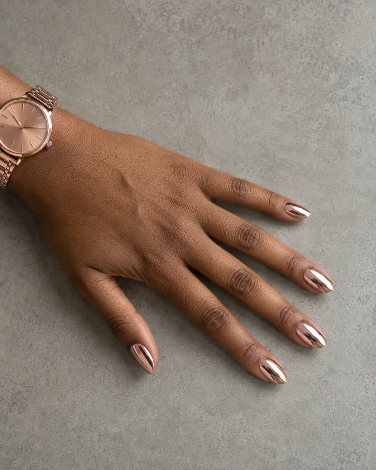 Single left hand with oval nails in mirror chrome rose gold with a rose gold watch band visible at wrist on soft grey concrete surface