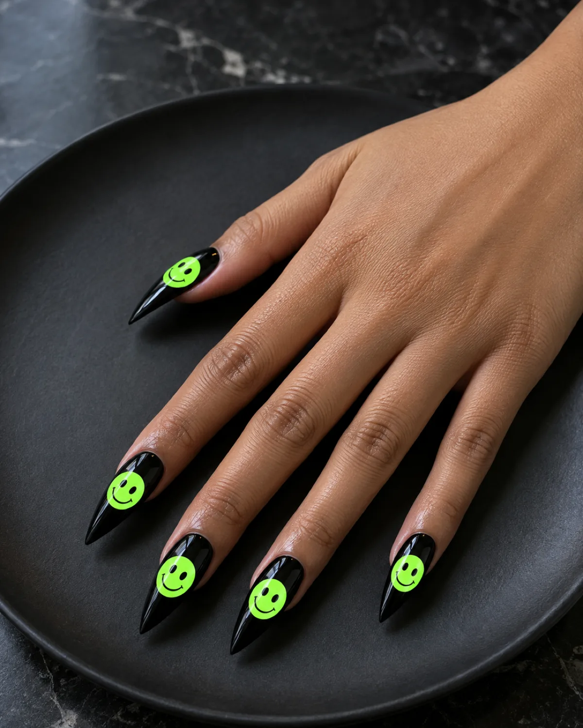Single right hand with neon green smiley face nails on long stiletto shape resting on a matte black ceramic plate