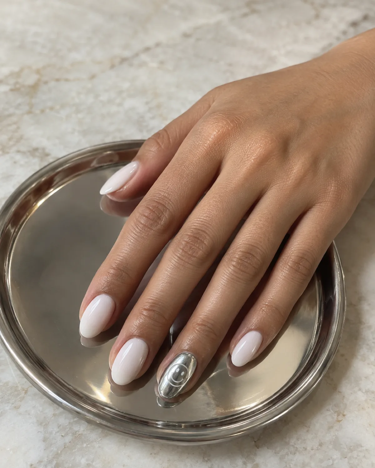 Single right hand with chrome smiley accent nail on short almond shape resting on a small round silver tray
