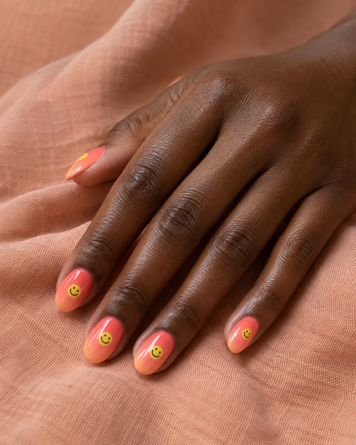 Single right hand with smiley face nails on coral-to-peach gradient on medium round shape resting on a peach-colored linen cloth