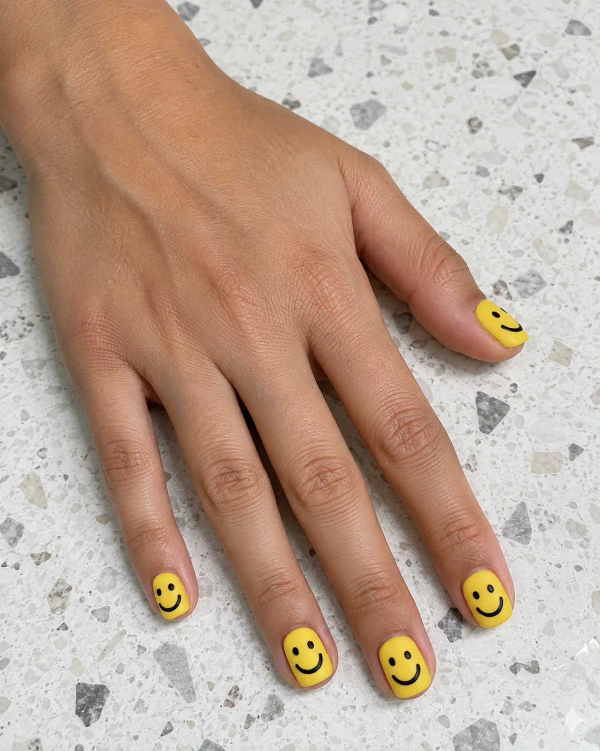 Single left hand with matte yellow full-smiley nails on short squoval shape resting on a white terrazzo surface