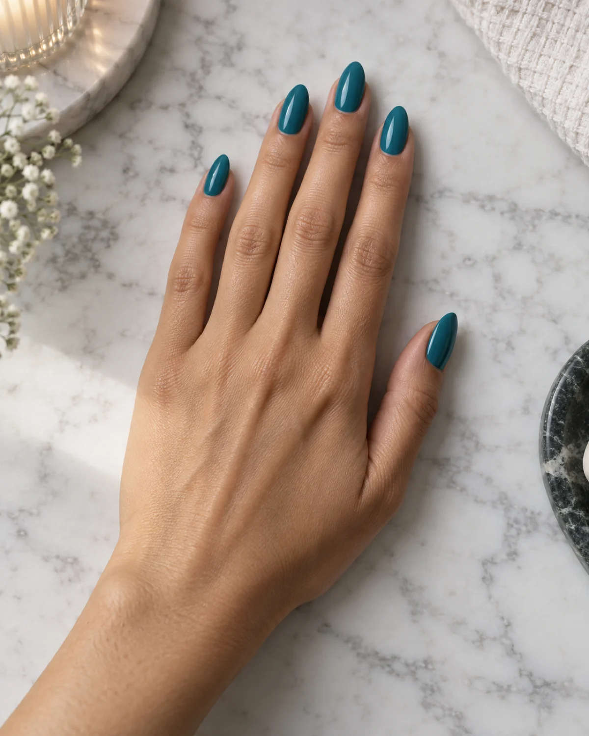 Single right hand with almond nails in rich mid-teal crème polish resting palm down on white marble countertop