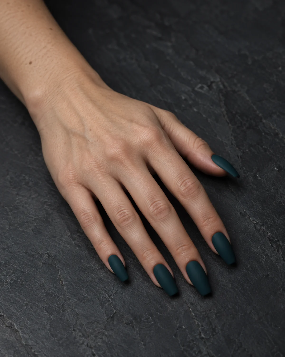 Single left hand with coffin nails in deep dark teal matte finish resting palm down on dark slate surface