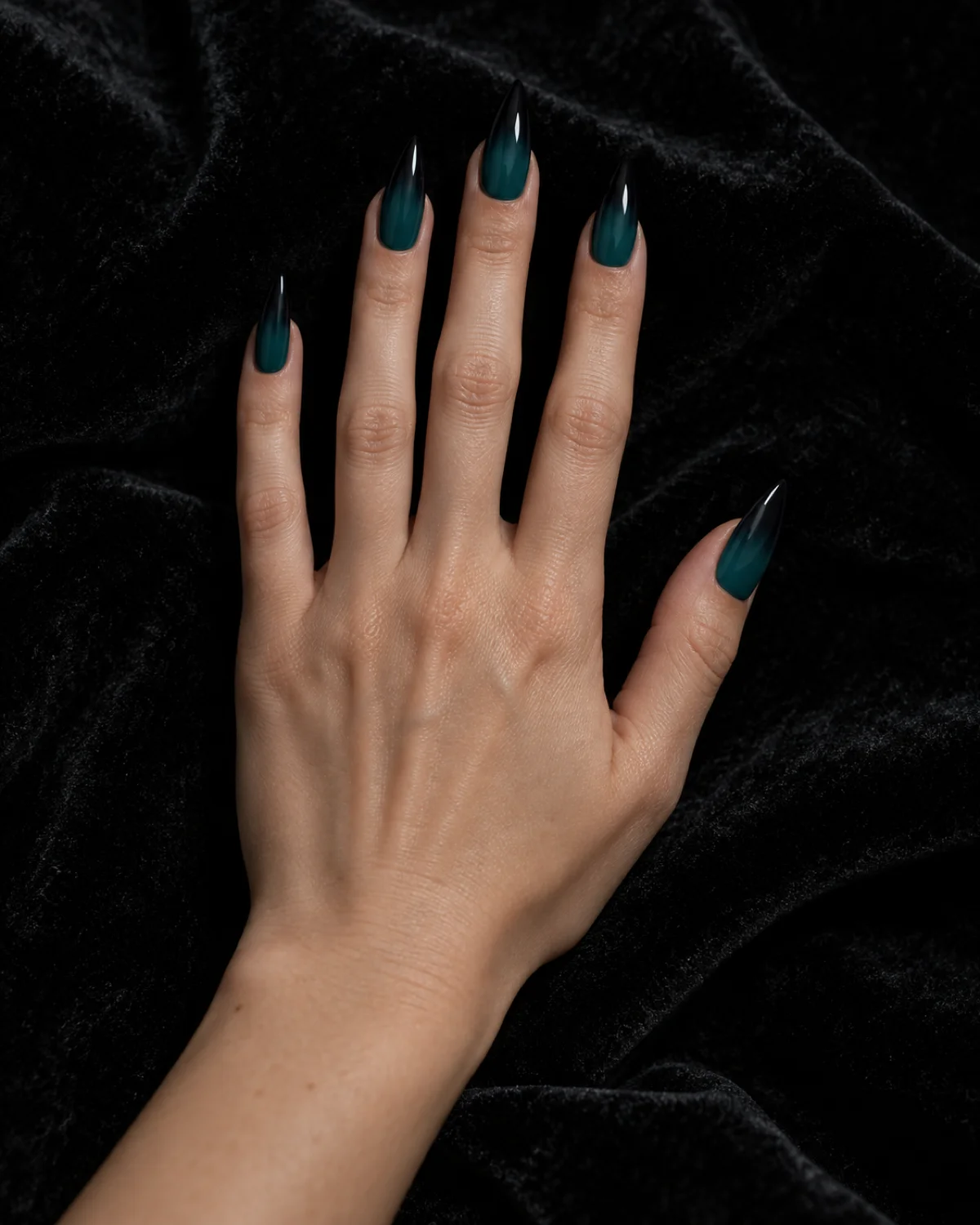 Single right hand with stiletto nails showing teal-to-black ombré gradient resting palm down on black velvet surface